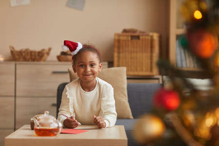 Portrait of cute African-American girl writing letter to Santa while sitting at table by Christmas tree, copy spaceの写真素材
