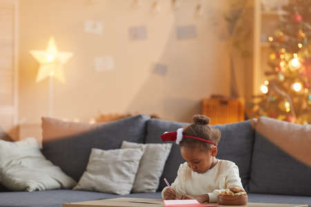 Portrait of cute African-American girl writing letter to Santa while sitting at table in cozy living room with Christmas tree in background, copy spaceの写真素材
