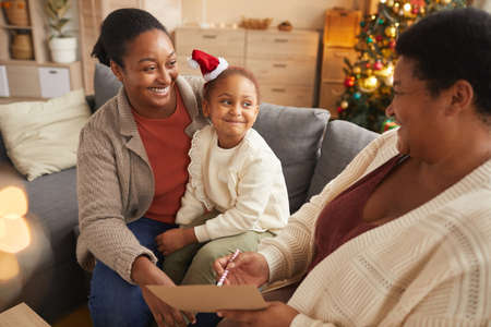 Warm-toned portrait of smiling African-American family writing letter to Santa with cute little girl while enjoying Christmas season at homeの写真素材