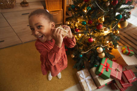 Fisheye portrait of excited African-American girl opening Christmas presents while standing by tree at home, copy spaceの写真素材