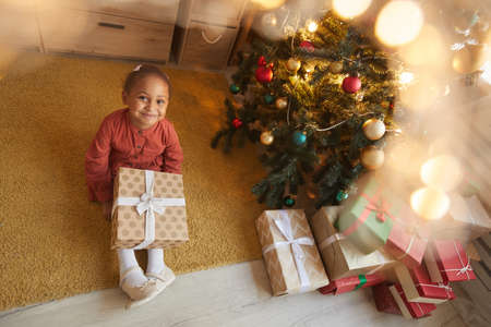 Above view portrait of happy African-American girl opening Christmas presents while sitting by tree...