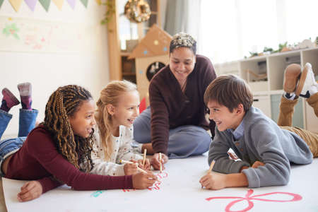 Full length portrait of smiling female teacher sitting on floor with multi-ethnic group of kids...