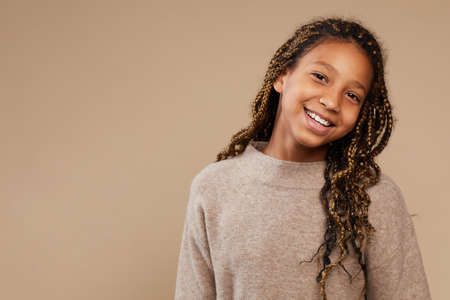 Portrait of carefree African-American girl smiling happily at camera while standing against beige background in studio, copy spaceの写真素材