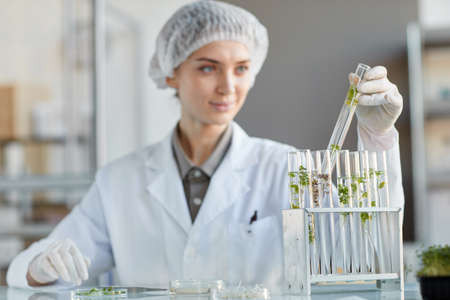 Portrait of young female scientist holding test tube with plant samples while working on research in...