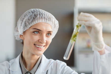 Close up portrait of smiling female scientist holding test tube with plant samples while working on...