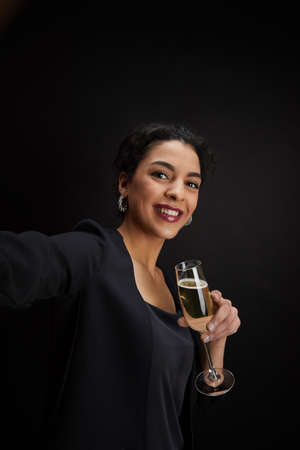 Vertical portrait of elegant Middle-Eastern woman holding champagne glass and taking selfie photo while standing against black background at party, copy spaceの写真素材