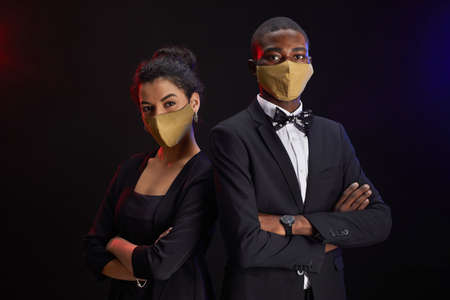 Waist up portrait of elegant mixed race couple wearing face masks while posing against black background at party, copy spaceの写真素材