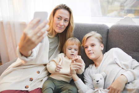 Portrait of modern mature mother taking selfie photo with two kids while relaxing together on sofa at home, copy spaceの写真素材