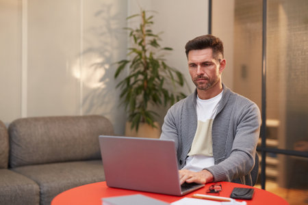 Portrait of handsome mature man working with laptop at red table in minimal office interior, copy spaceの写真素材
