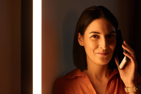 Minimal close up portrait of smiling elegant woman speaking by smartphone and looking at camera lit...