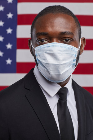 Vertical portrait of African-American politician wearing mask and looking at camera while standing against USA flag backgroundの写真素材