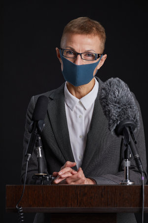 Vertical portrait of mature woman wearing mask while giving speech standing at podium against black backgroundの写真素材