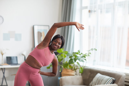 Portrait of smiling African-American woman wearing pink sportswear while stretching during workout...