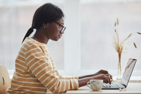 Side view portrait of young African-American woman using laptop while sitting at table in cozy home interiorの写真素材