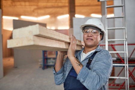 Waist up portrait of smiling female worker carrying wood boards while working on construction site, copy spaceの写真素材
