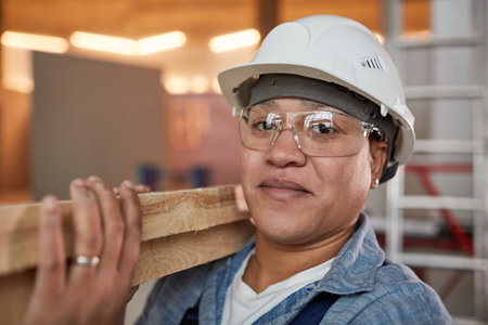 Close up portrait of smiling female worker carrying wood boards while working on construction site, copy spaceの写真素材