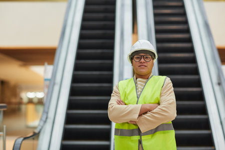 Graphic waist up portrait of mixed-race female worker looking at camera while standing with arms crossed against escalator in business mall, copy spaceの写真素材