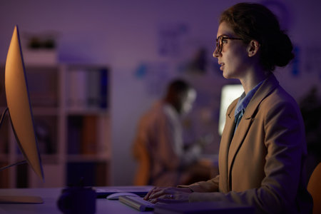 Side view shot portrait of serious young woman wearing eyeglasses sitting in front of computer monitor working in darknessの写真素材