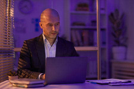 Serious mature man sitting at office desk working on business plan using laptop late in evening,...