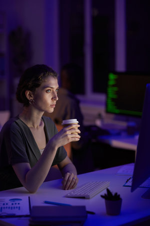 Modern young woman sitting in dark office room late in evening drinking coffee looking at computer monitorの写真素材