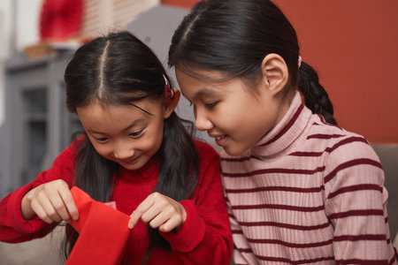 Medium close-up portrait of two curious little Chinese girls looking into red envelope, Lunar New...