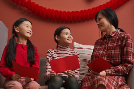 Modern Chinese woman spending time at home with her little daughters preparing red envelopes for...