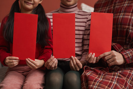 Medium section shot of Asian family of three sitting in sofa holding handmade red envelopes for Chinese New Year giftsの写真素材