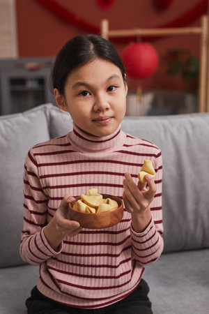 Medium portrait of young Asian girl sitting on sofa in living room choosing fortune cookie looking at cameraの写真素材