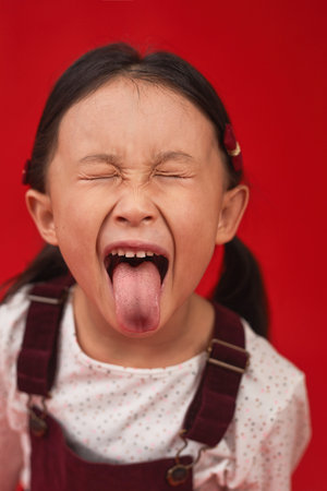 Close-up portrait of funny little Asian girl with two ponytails grimacing on camera sticking with...