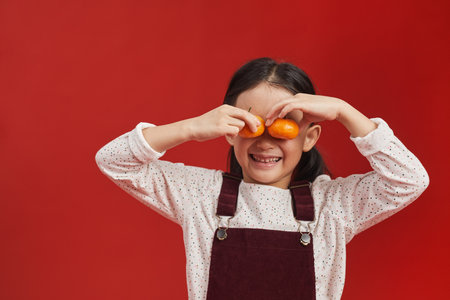 Studio portrait shot of cute little Asian girl standing against red wall background covering eyes with tangerinesの写真素材