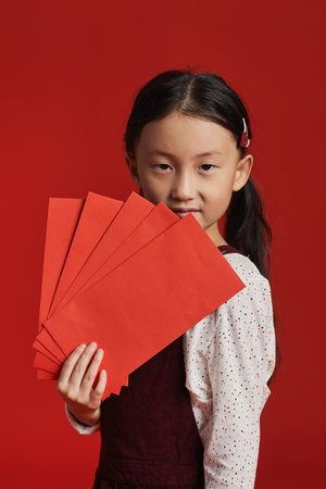 Vertical medium studio portrait of cute Asian girl with two ponytails holding bunch of red envelopes, red backgroundの写真素材