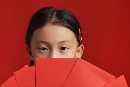 Close-up studio eyes only portrait of cute Asian girl with two holding bunch of red envelopes in front of her face, red backgroundの写真素材