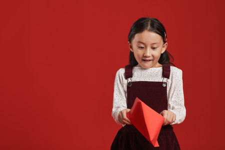 Medium studio portrait of cheerful little Chinese girl standing against red wall background looking into red envelopeの写真素材