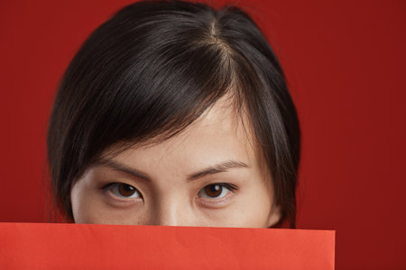 Close-up studio shot of unrecognizable Asian woman covering her face with red envelope looking at cameraの写真素材