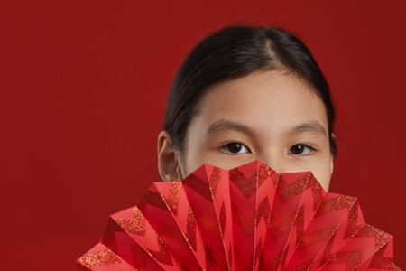 Pretty Asian girl with beautiful eyes hiding her face behind red fan looking at camera, red backgroundの写真素材