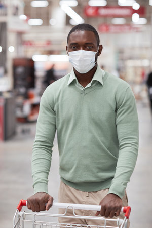 Vertical portrait of African-American man wearing mask in supermarket and looking at camera while pushing shopping cartの写真素材