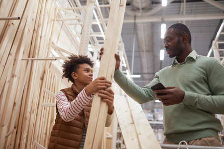 Low angle portrait of African-American father and son shopping together in hardware store, focus on man choosing wooden boards for construction or home improvementの写真素材