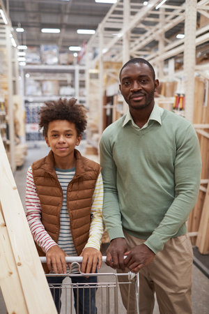 Vertical portrait of African-American father and son shopping together in hardware store both looking at camera while pushing cart with wooden boardsの写真素材