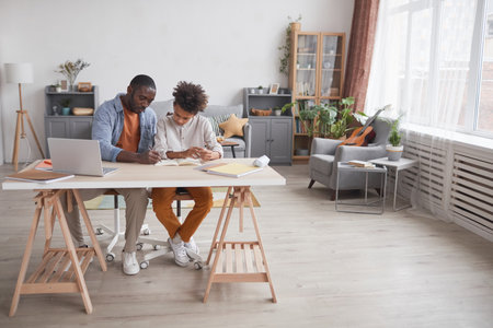 Full length portrait of caring African-American father helping son doing homework or studying while sitting together at desk in home interior, copy spaceの写真素材