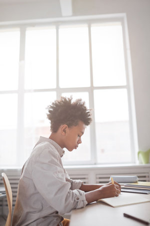Vertical side view portrait of teenage African-American boy doing homework while sitting at desk...