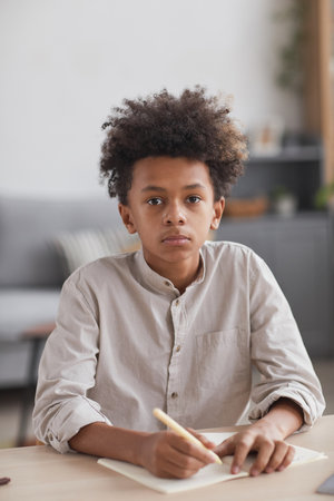 Vertical front view portrait of teenage African-American boy doing homework at desk and looking at camera in modern home interiorの写真素材