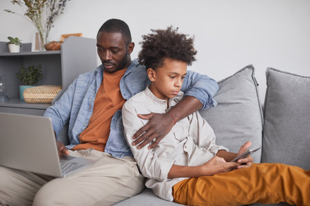 African American man and his teen son sitting together on sofa in living room surfing Internet using laptop and smartphoneの写真素材