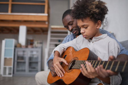 Mature man spending spare time with his son at home, teaching him how to play beautiful song on guitarの写真素材
