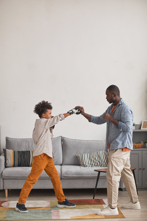 Modern African American father practicing boxing punches with his teen son in living room at home, vertical side view long shotの写真素材