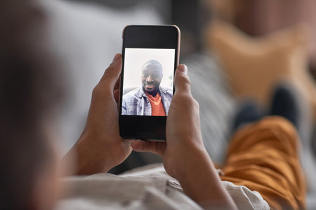 Unrecognizable child relaxing on sofa holding smartphone in his hands having online call with his fatherの写真素材
