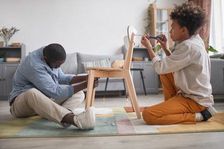 Modern African American man spending time with his teen son in living room at home assembling new chairの写真素材