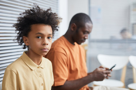 Portrait of teenage African-American boy looking at camera while waiting in line at medical clinic with father, copy spaceの写真素材