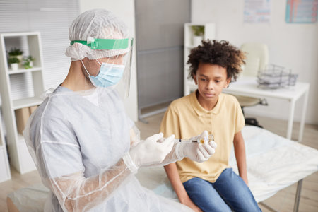 High angle portrait of African-American boy looking at nurse drawing medicine into syringe during...
