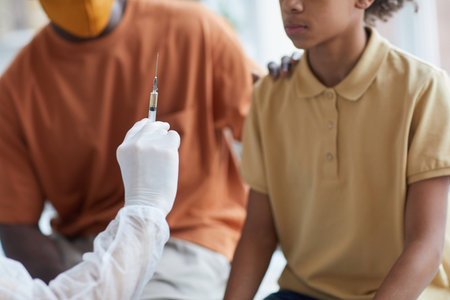 Close up of African-American teenage boy looking at nurse holding syringe with needle during covid vaccination in clinic with father supporting himの写真素材