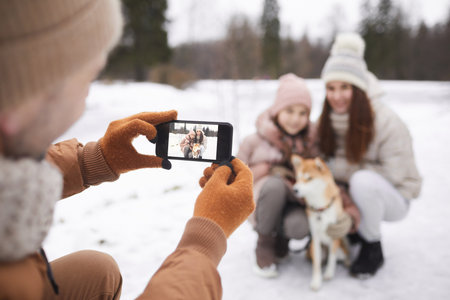 Portrait of father taking photo of cute daughter and wife with dog while enjoying walk outdoors together in winter forest, focus on smartphone screen, copy spaceの写真素材